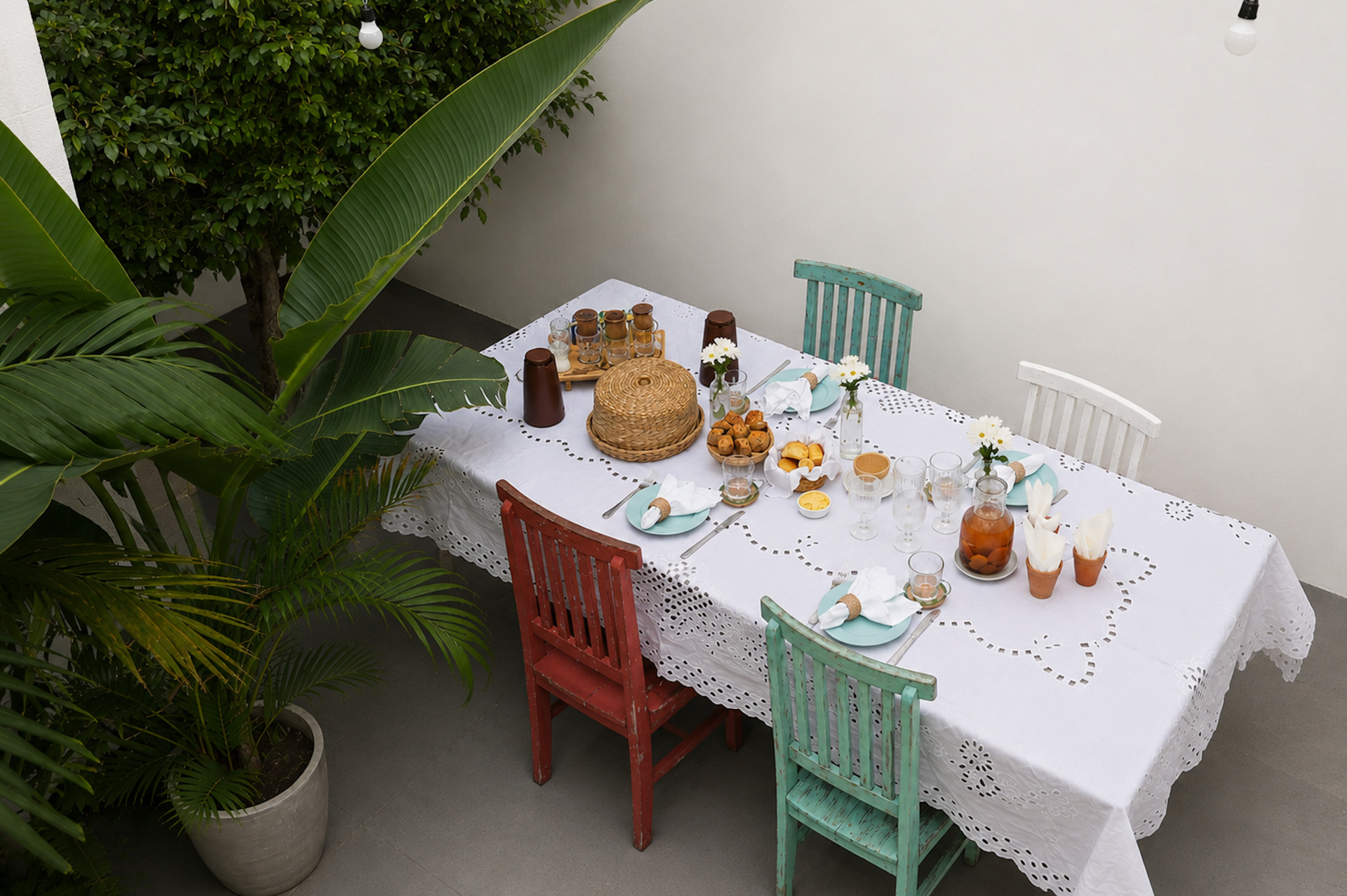 Mesa posta vista de cima, com toalha branca, jarras e cadeiras coloridas, cercada de plantas